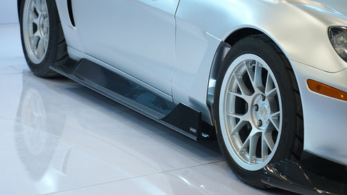 Close-up of a silver wide-body Chevrolet Corvette performance car displayed indoors, featuring aggressive aerodynamic bodywork, carbon fiber front splitter, vented hood, and lightweight multi-spoke wheels. The image highlights the custom motorsport styling and premium carbon fiber aero components under exhibition lighting.