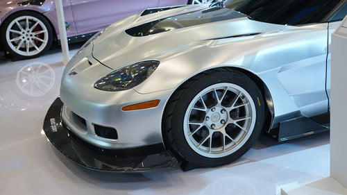 Close-up of a silver wide-body Chevrolet Corvette performance car displayed indoors, featuring aggressive aerodynamic bodywork, carbon fiber front splitter, vented hood, and lightweight multi-spoke wheels. The image highlights the custom motorsport styling and premium carbon fiber aero components under exhibition lighting.