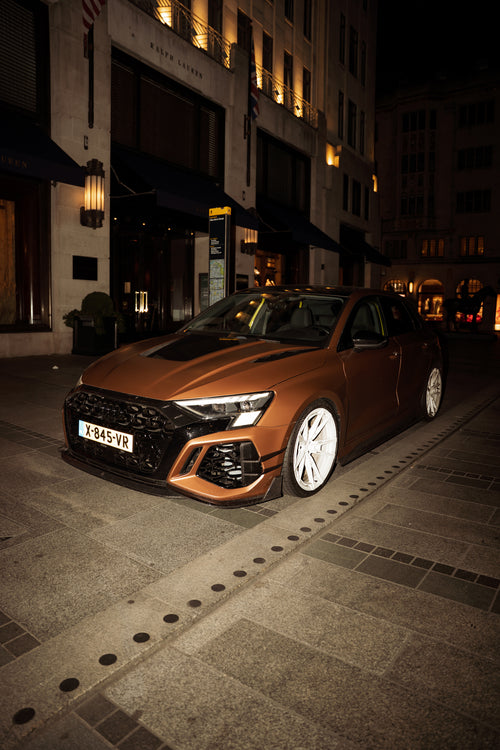 Bronze Audi RS4 Avant featuring a REVOZPORT carbon fiber body kit with front lip and side skirts, paired with silver multi-spoke forged wheels, parked in a modern urban setting.