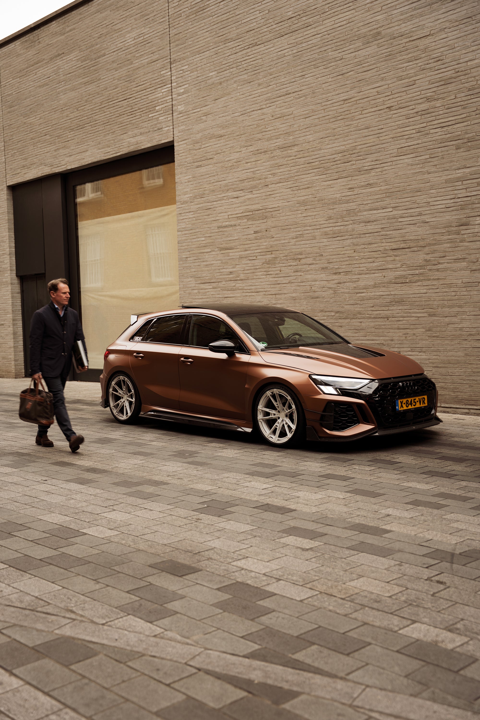 Bronze Audi RS4 Avant featuring a REVOZPORT carbon fiber body kit with front lip and side skirts, paired with silver multi-spoke forged wheels, parked in a modern urban setting.
