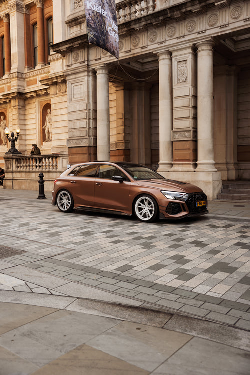 Bronze Audi RS4 Avant featuring a REVOZPORT carbon fiber body kit with front lip and side skirts, paired with silver multi-spoke forged wheels, parked in a modern urban setting.