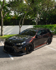 BMW touring car with carbon fiber body kit parked in a leafy outdoor lot, highlighting front lip and lowered stance.
