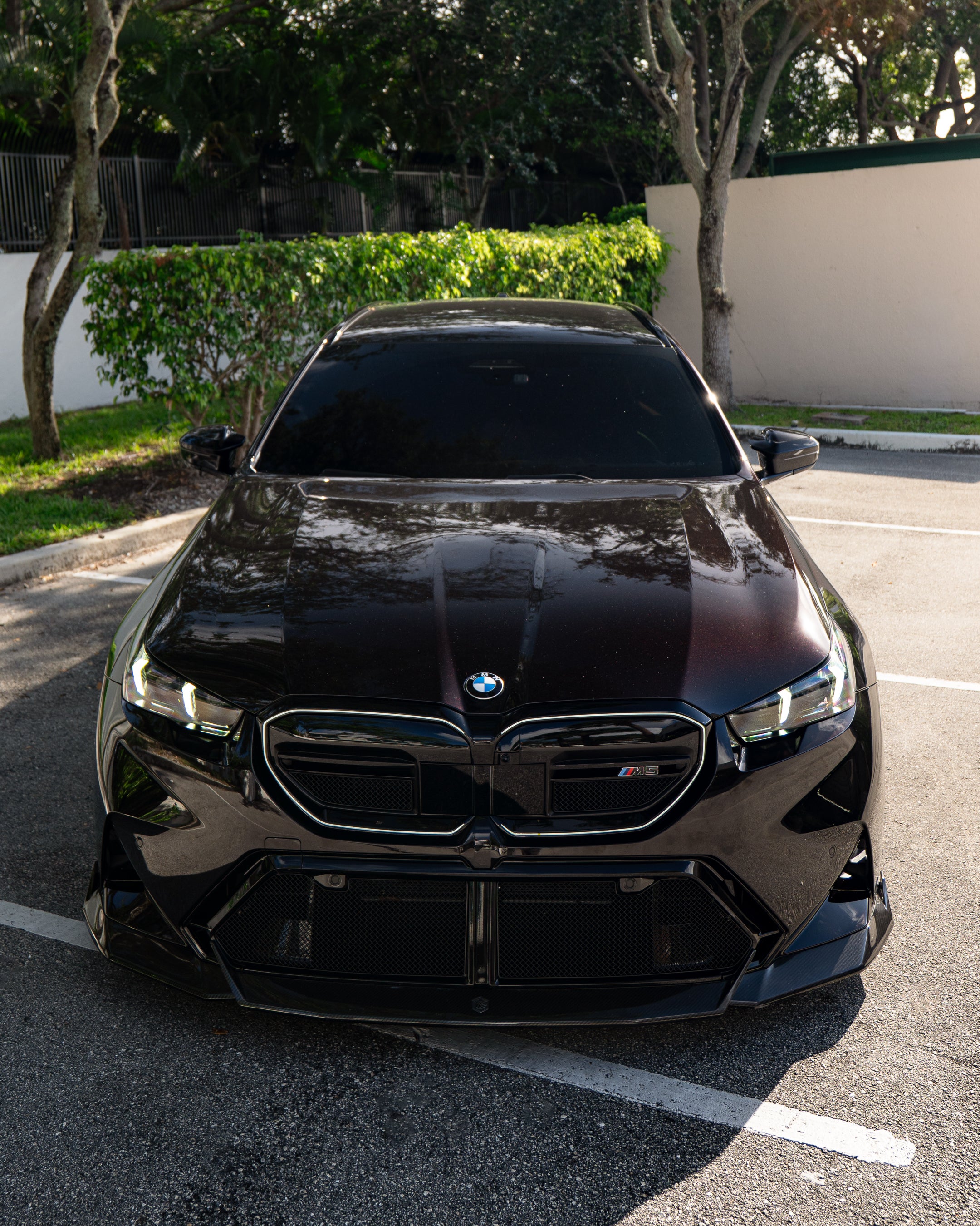 BMW touring car with carbon fiber body kit parked in a leafy outdoor lot, highlighting front lip and lowered stance.
