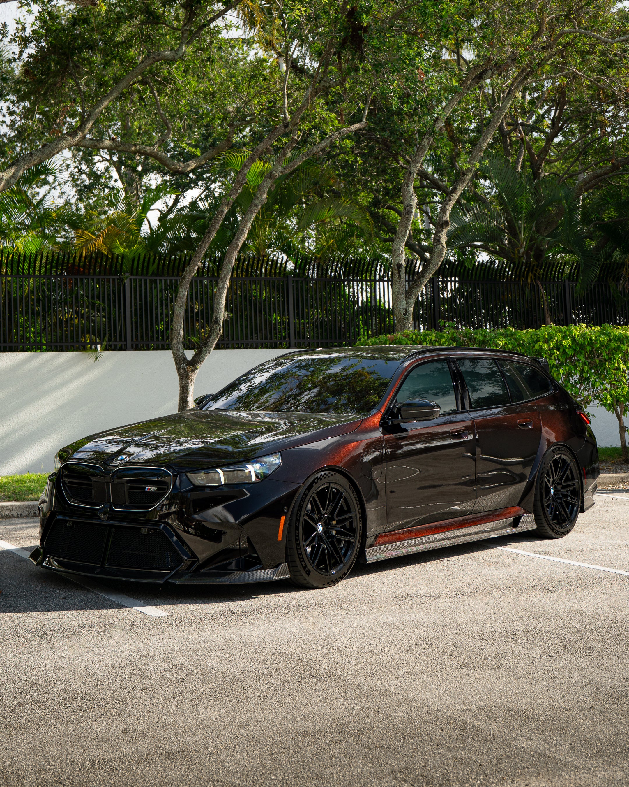BMW touring car with carbon fiber body kit parked in a leafy outdoor lot, highlighting front lip and lowered stance.