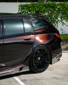 Close-up of a BMW rear wheel with carbon fiber side skirt and diffuser detail, highlighting glossy weave and performance styling.