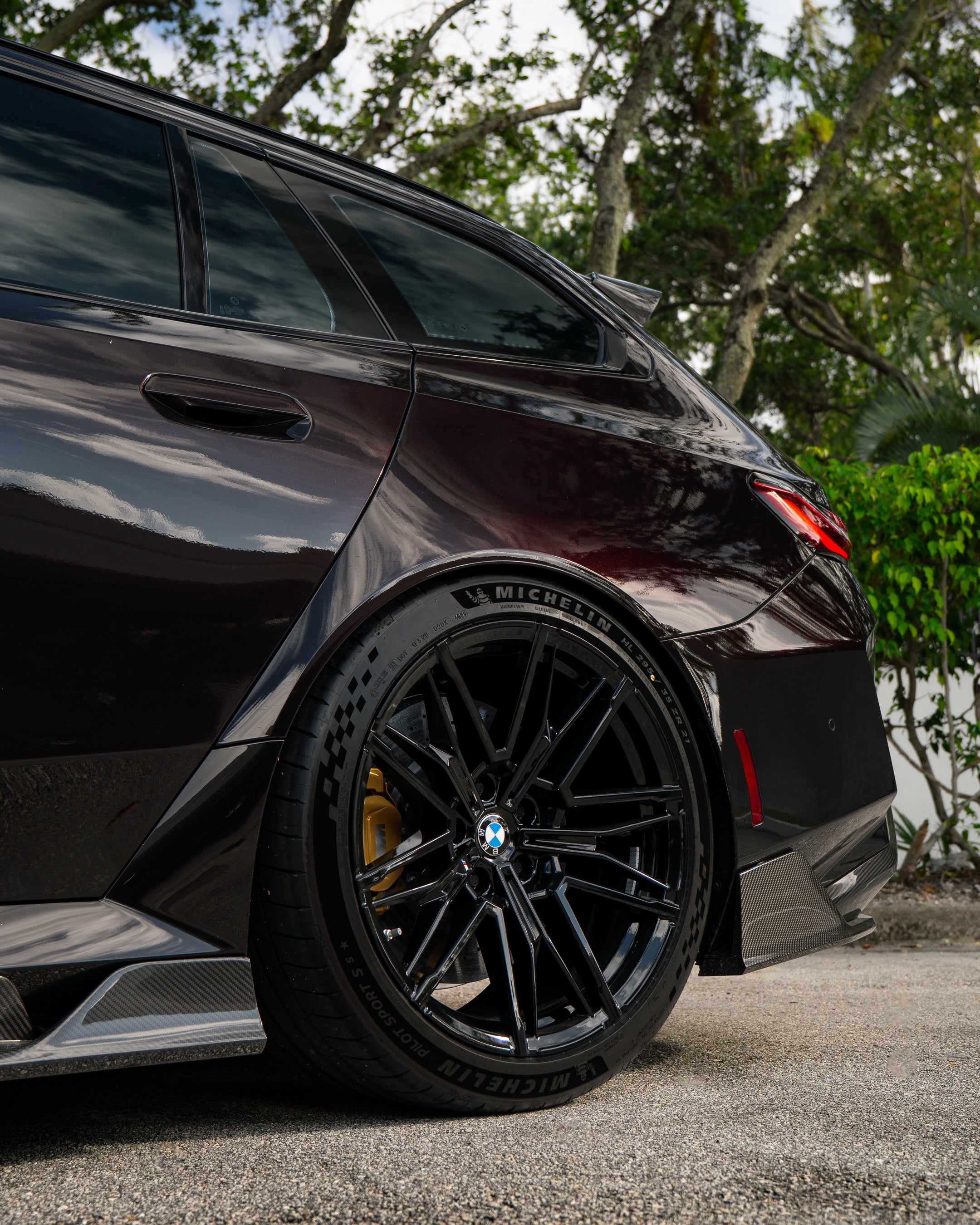 Close-up of a BMW rear wheel with carbon fiber side skirt and diffuser detail, highlighting glossy weave and performance styling.