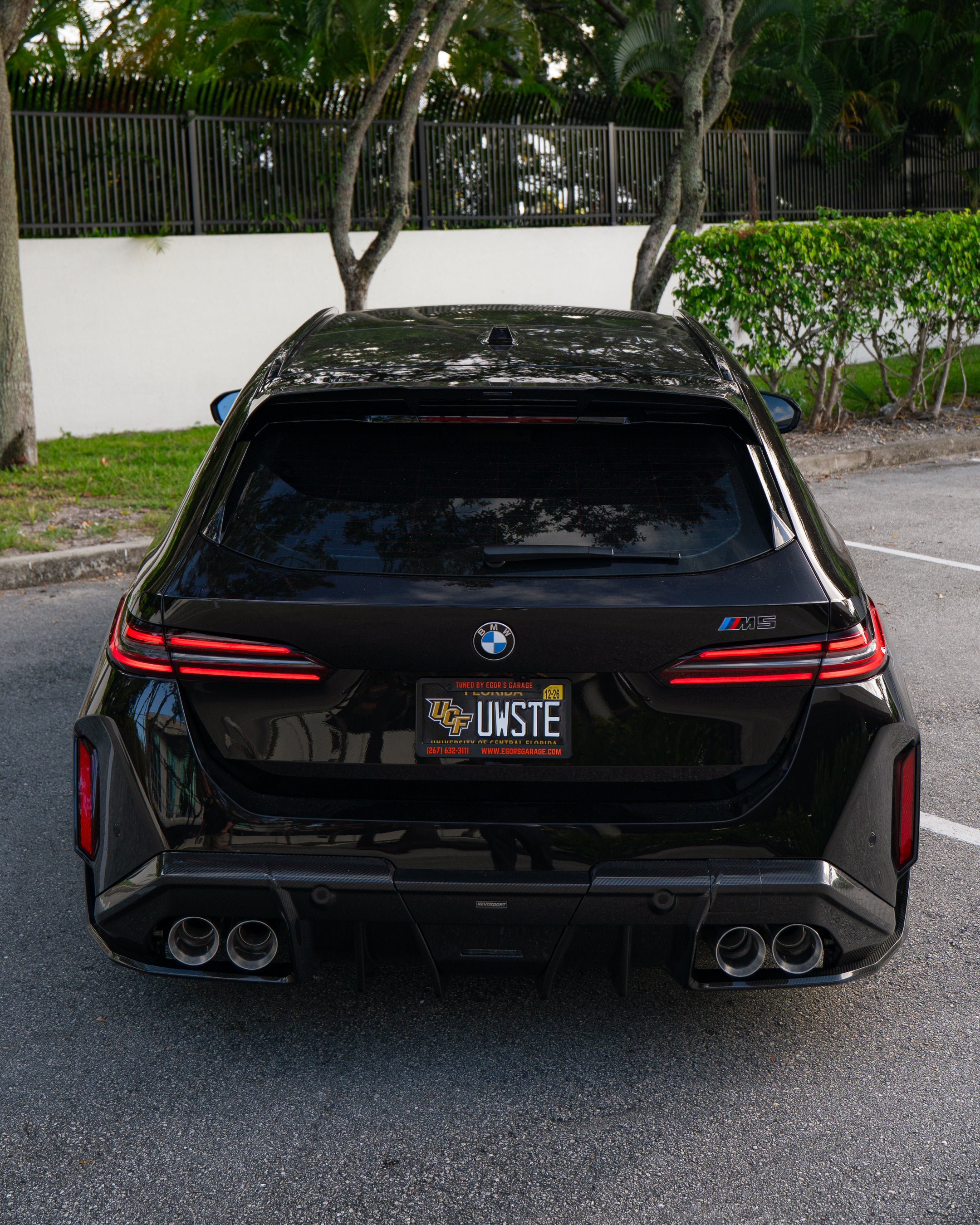 Rear three-quarter view of a BMW touring car with carbon fiber aero kit, rear diffuser, roof spoiler, and quad exhausts parked in a leafy outdoor setting.