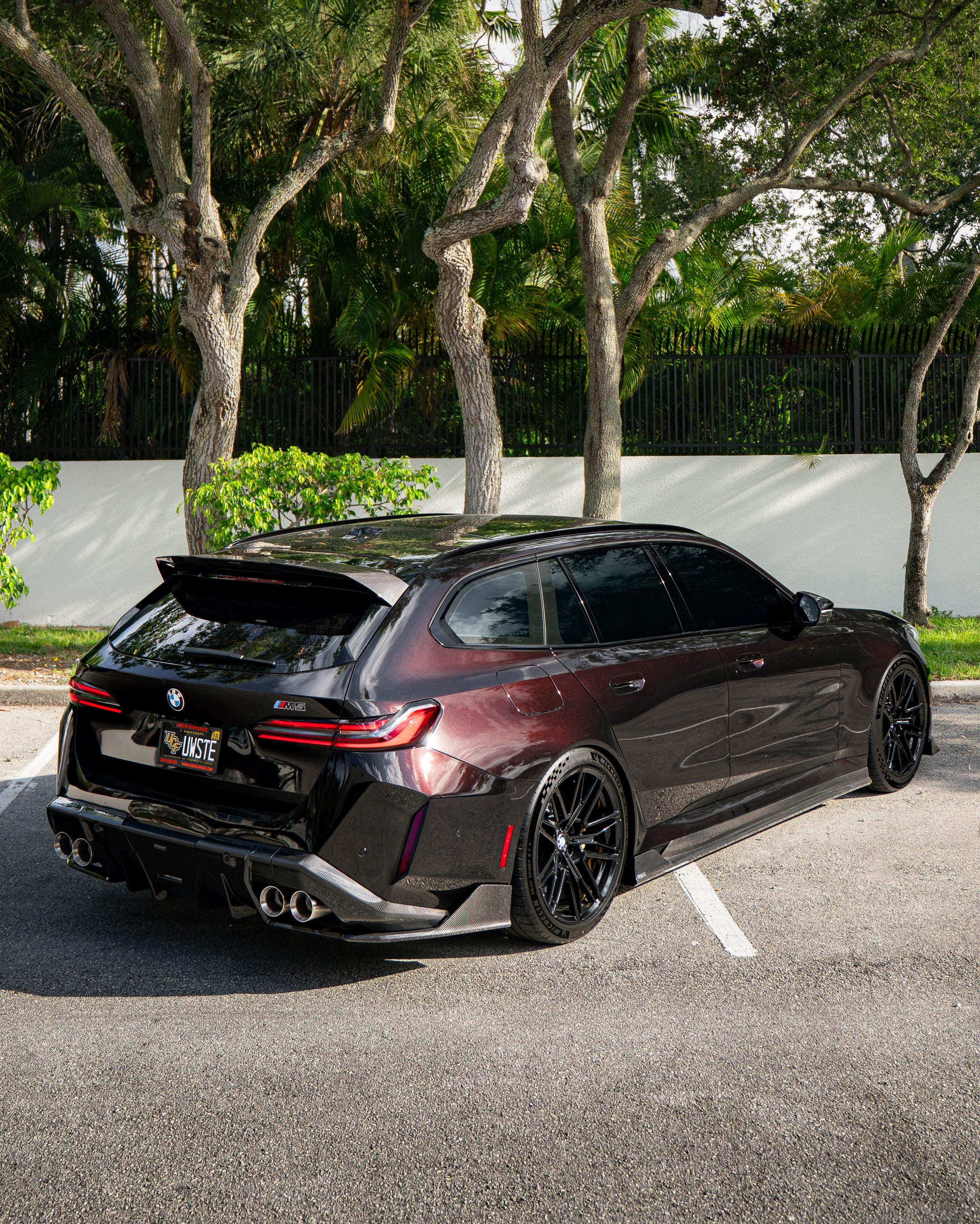 Rear three-quarter view of a BMW touring car with carbon fiber aero kit, rear diffuser, roof spoiler, and quad exhausts parked in a leafy outdoor setting.