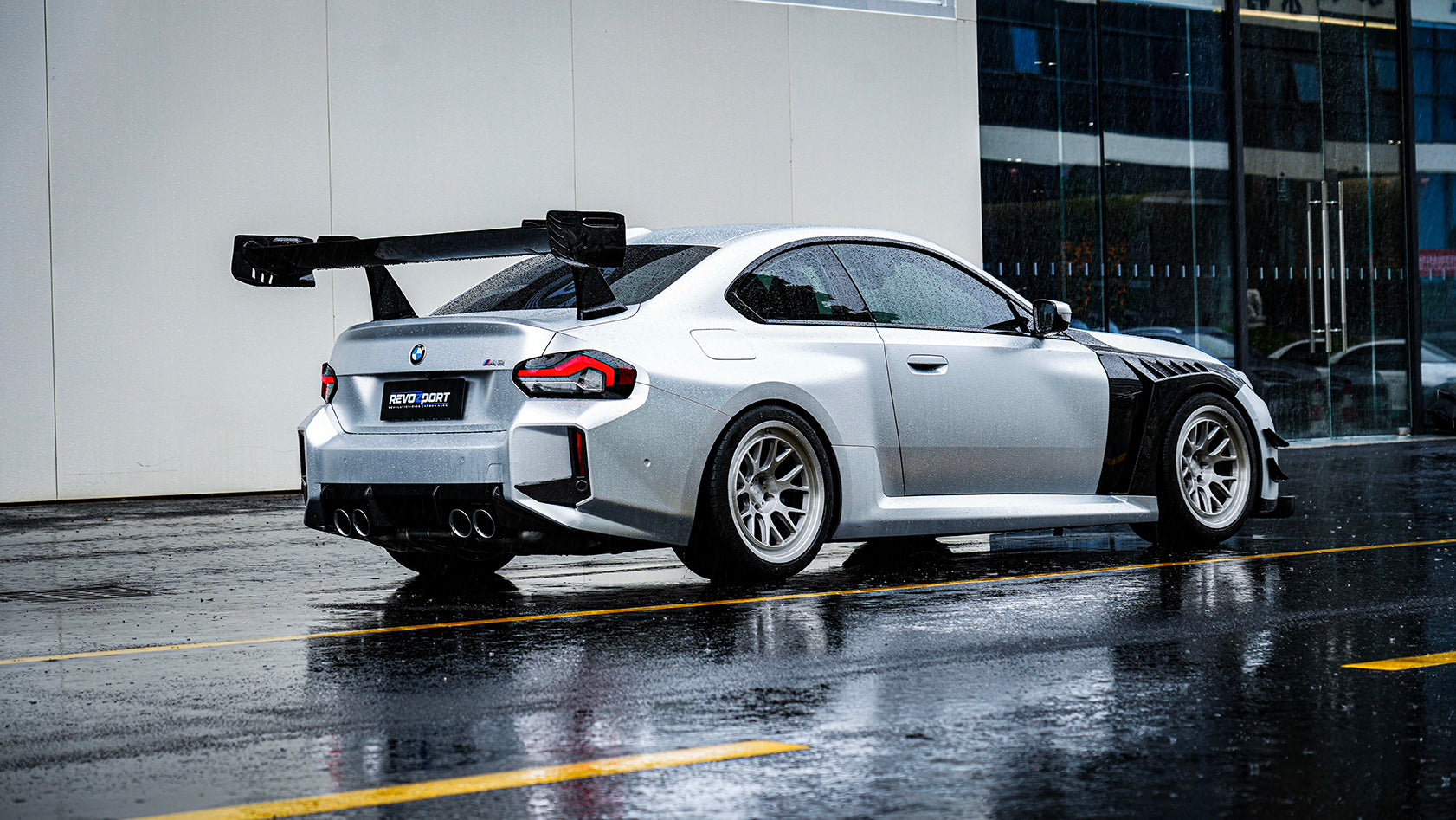 White BMW G82 M4 with carbon fiber hood, vented fenders, side skirts, and oversized GT wing driving through heavy rain on a wet urban road.