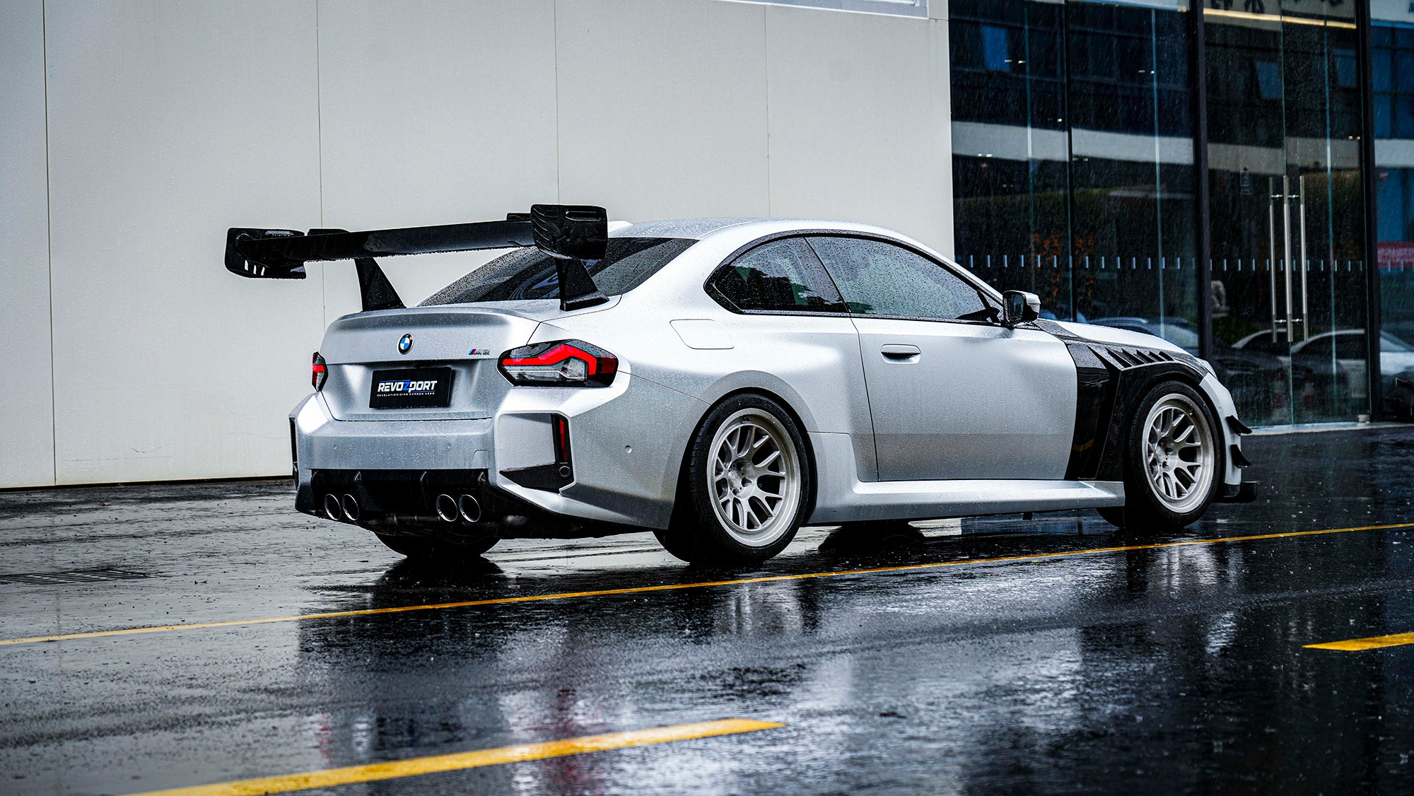Rear three-quarter view of a BMW with carbon fiber widebody kit, massive rear wing, white wheels, and aggressive aero styling in rain.