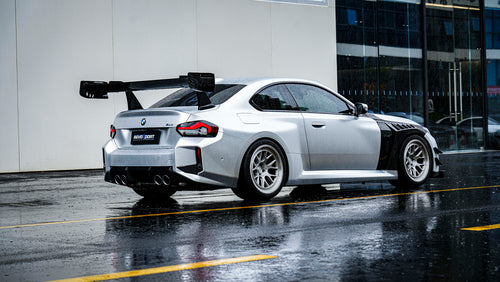 Rear three-quarter view of a BMW with carbon fiber widebody kit, massive rear wing, white wheels, and aggressive aero styling in rain.
