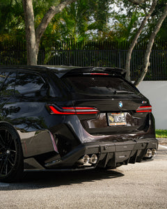 Rear three-quarter view of a BMW touring car with carbon fiber aero kit, rear diffuser, roof spoiler, and quad exhausts parked in a leafy outdoor setting.