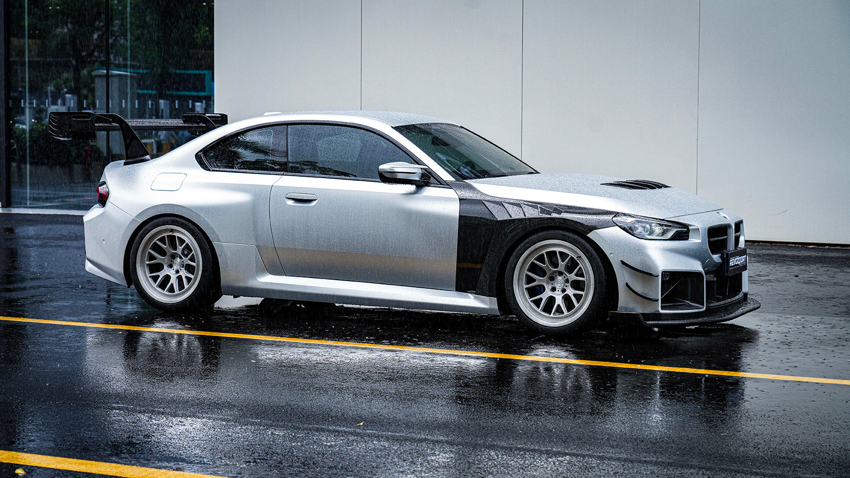 White BMW G82 M4 with carbon fiber hood, vented fenders, side skirts, and oversized GT wing driving through heavy rain on a wet urban road.
