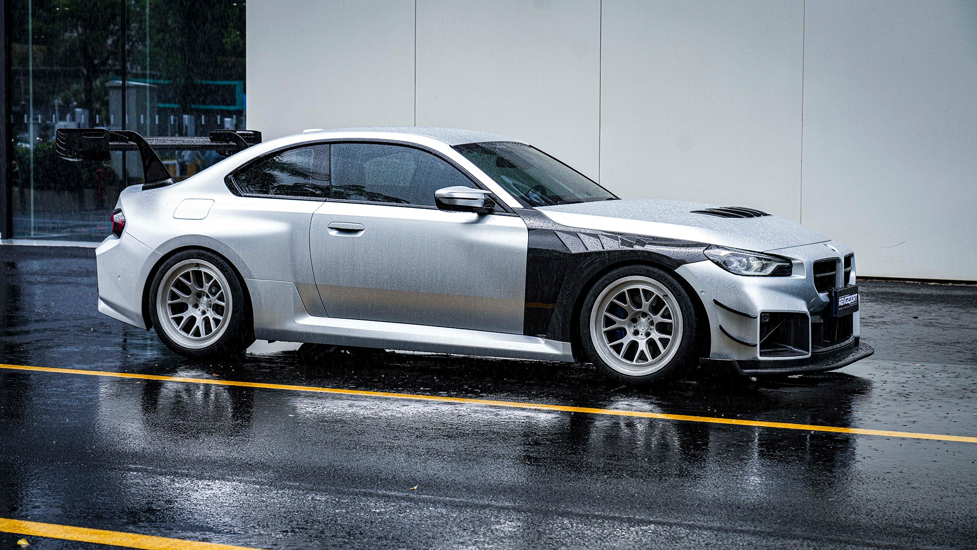Side profile of a BMW with carbon fiber widebody panels, oversized rear wing, track-inspired aero details, and rain reflections on wet pavement.
