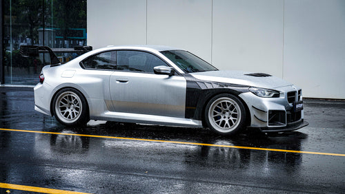 Side profile of a BMW with carbon fiber widebody panels, oversized rear wing, track-inspired aero details, and rain reflections on wet pavement.