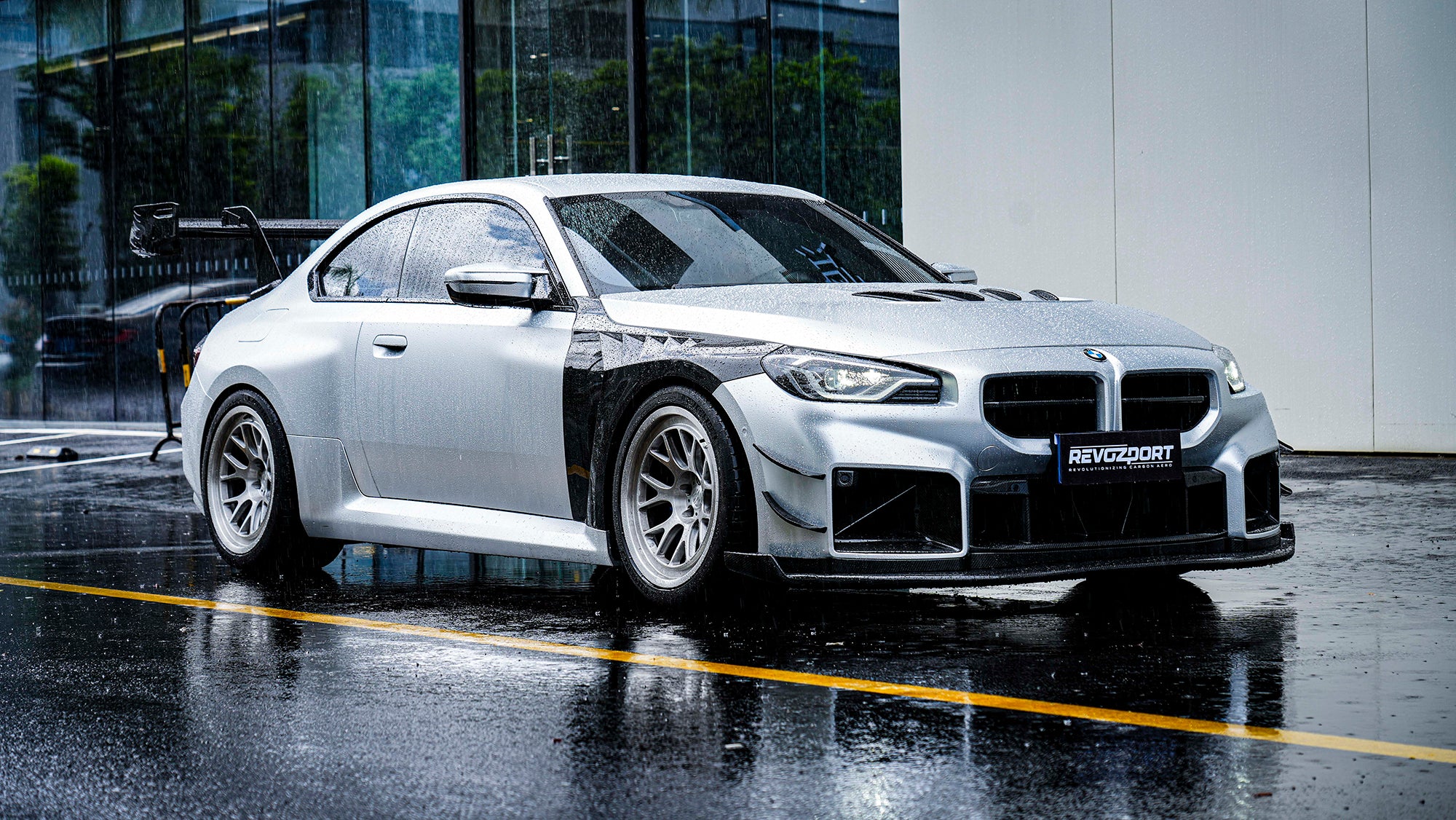 Front three-quarter view of a BMW featuring aggressive carbon fiber widebody aero, oversized rear wing, motorsport wheels, and rain-soaked street reflections.