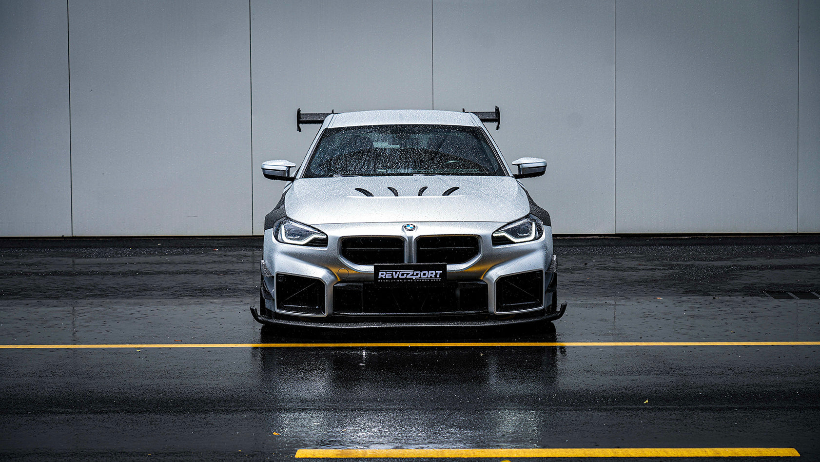 White BMW G82 M4 with carbon fiber hood, vented fenders, side skirts, and oversized GT wing driving through heavy rain on a wet urban road.