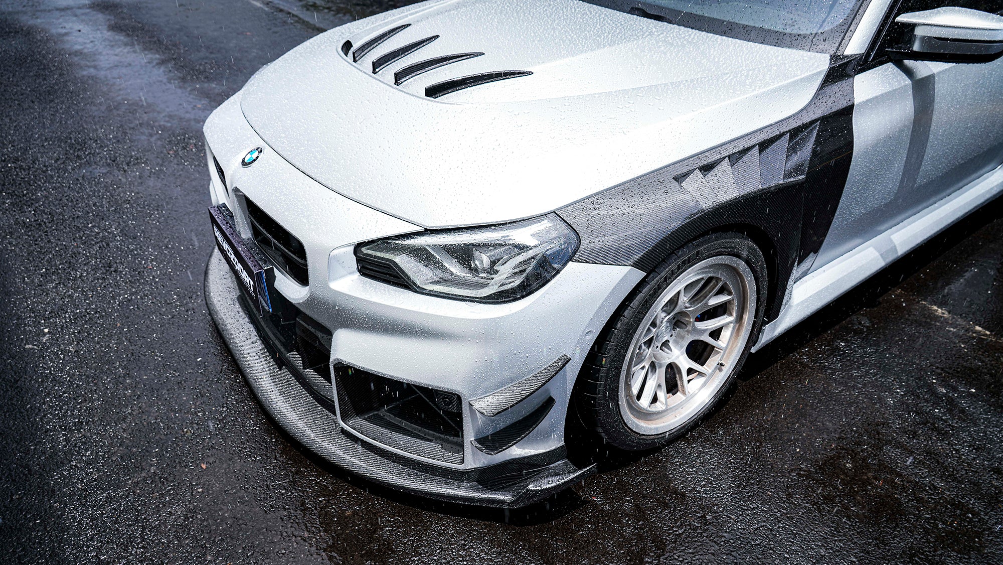 Close-up of a BMW with carbon fiber widebody kit, front splitter, vented hood, and white wheels on a rainy street.