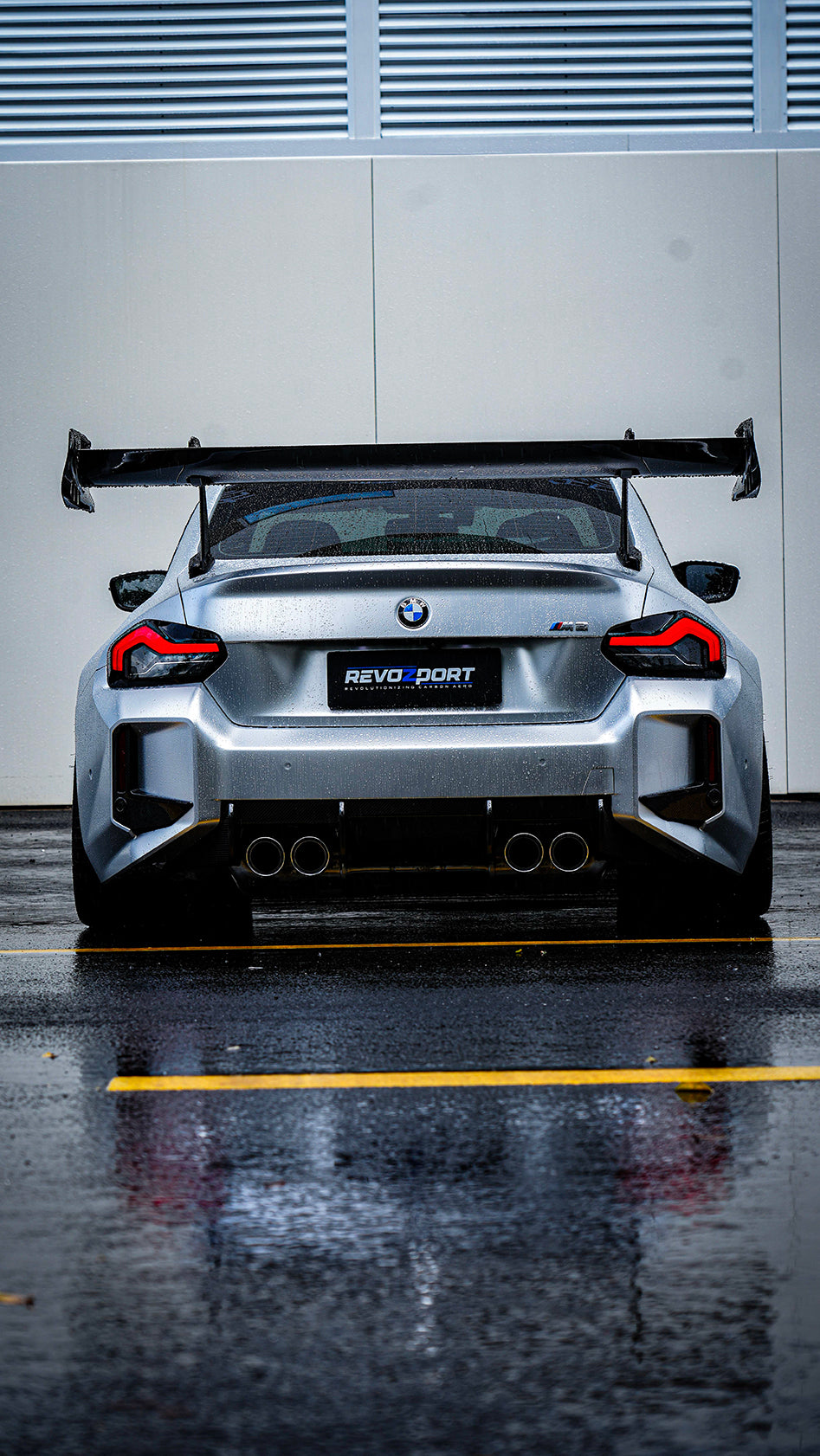 White BMW G82 M4 with carbon fiber hood, vented fenders, side skirts, and oversized GT wing driving through heavy rain on a wet urban road.