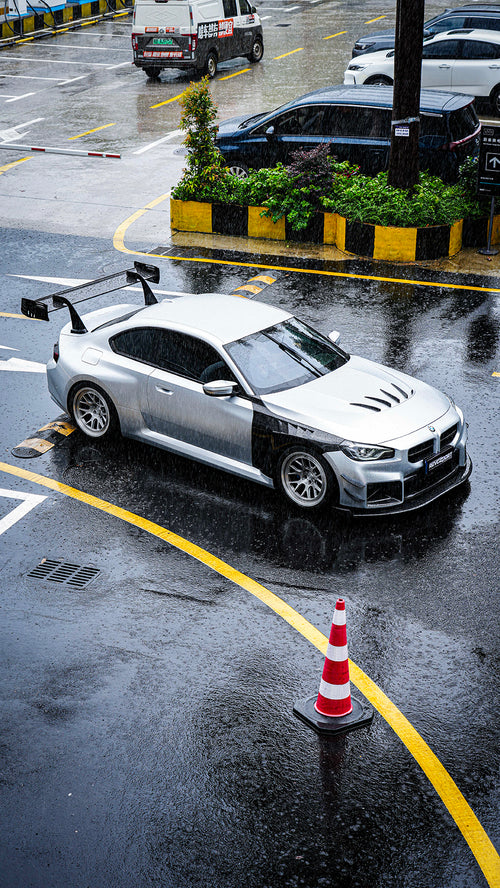White BMW G82 M4 with carbon fiber hood, vented fenders, side skirts, and oversized GT wing driving through heavy rain on a wet urban road.