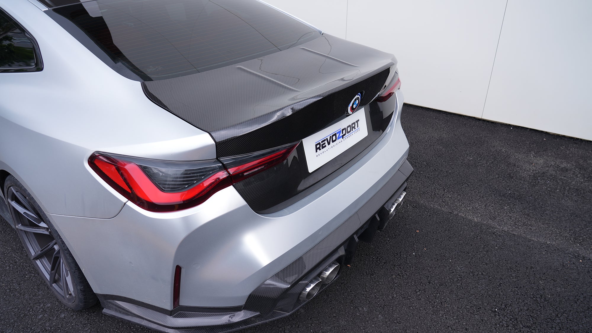 Close-up of a BMW G82 M4 rear end featuring a REVOZPORT carbon fiber trunk lid and integrated carbon rear spoiler, highlighting the glossy carbon weave detail.