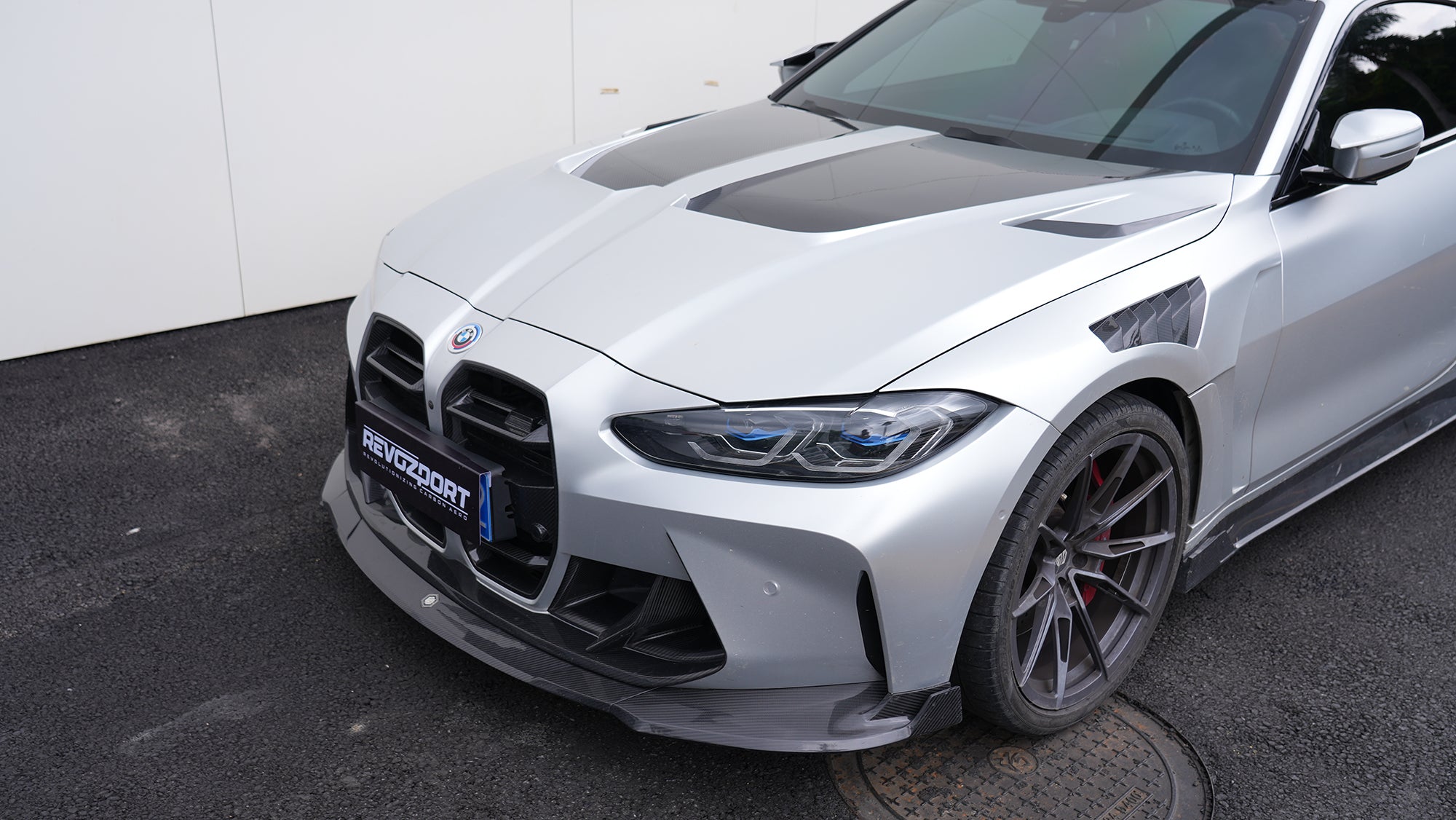 Silver BMW G82 M4 featuring a REVOZPORT carbon fiber body kit with front lip, side skirts, vented fenders, and aggressive aero styling parked outside a modern building.