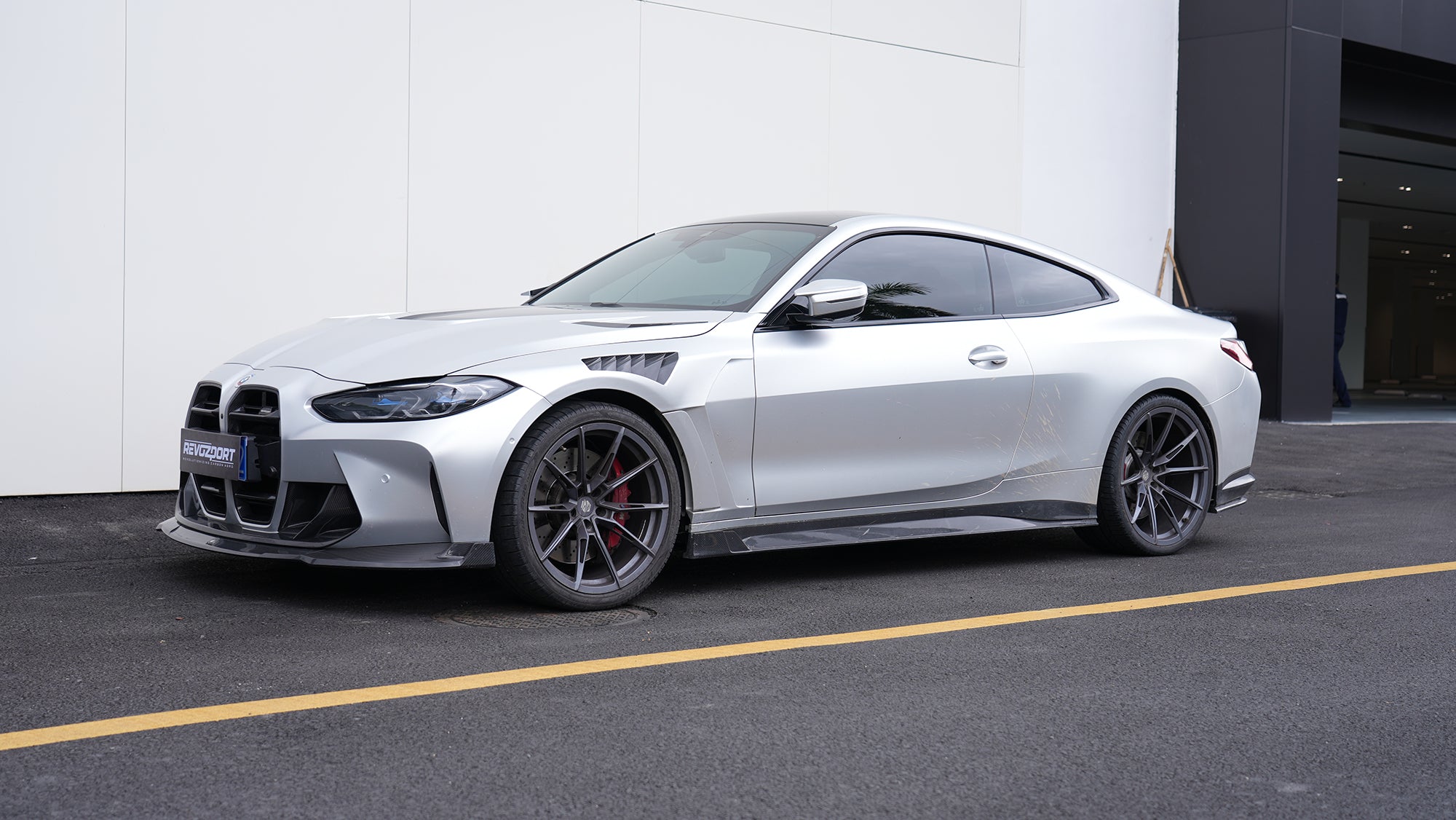 Silver BMW G82 M4 featuring a REVOZPORT carbon fiber body kit with front lip, side skirts, vented fenders, and aggressive aero styling parked outside a modern building.