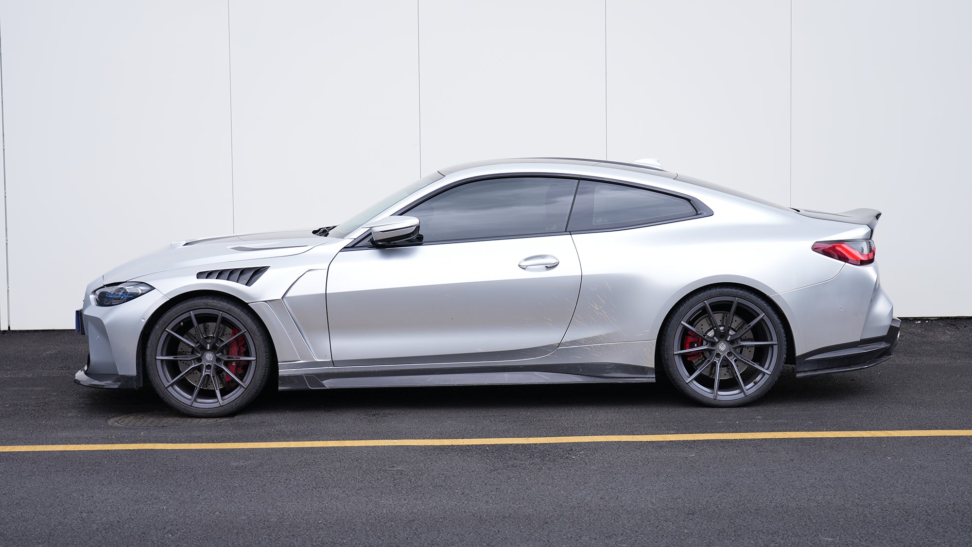 Silver BMW G82 M4 featuring a REVOZPORT carbon fiber body kit with front lip, side skirts, vented fenders, and aggressive aero styling parked outside a modern building.