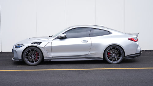 Silver BMW G82 M4 featuring a REVOZPORT carbon fiber body kit with front lip, side skirts, vented fenders, and aggressive aero styling parked outside a modern building.