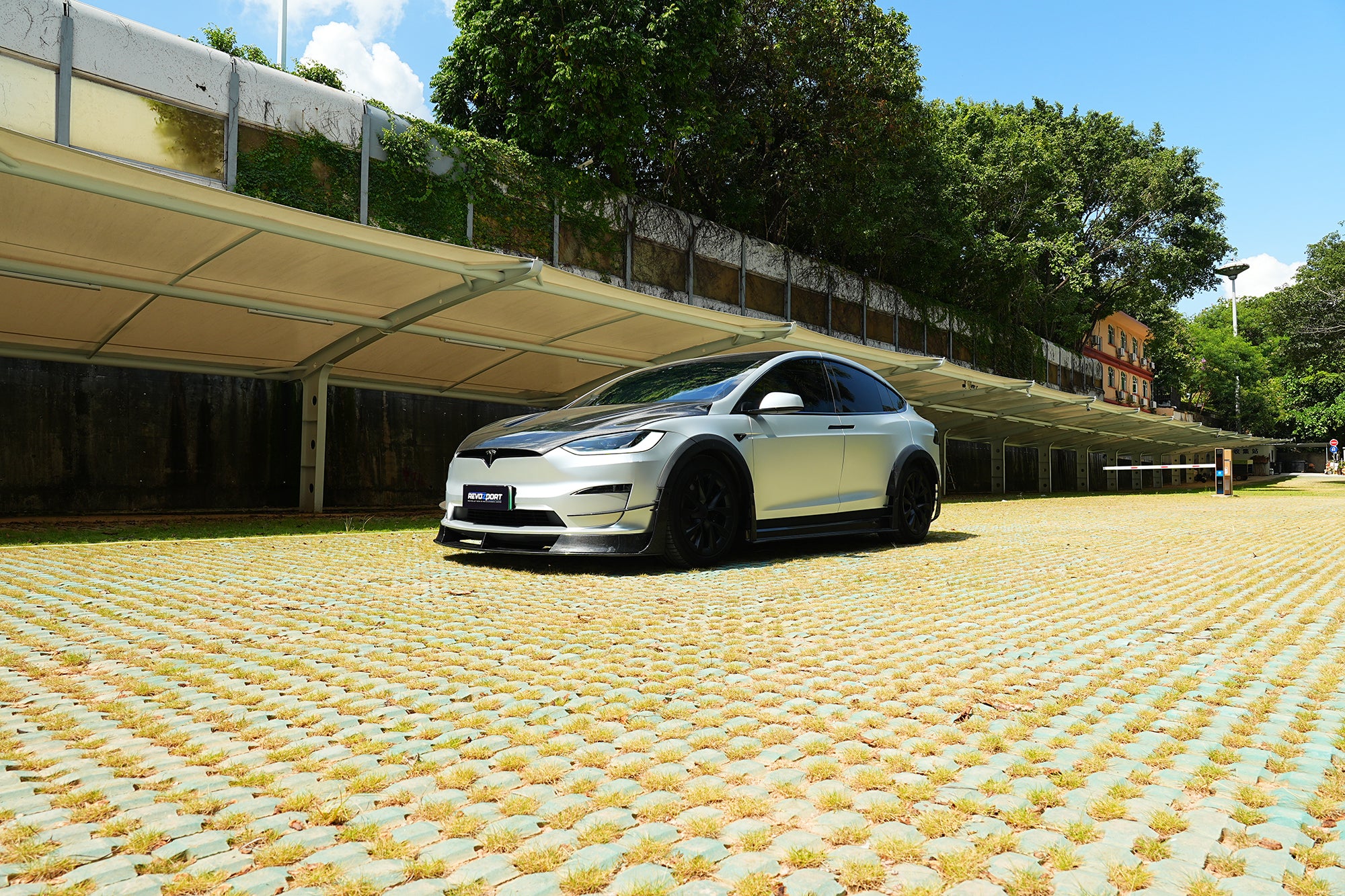 Silver Tesla Model X featuring a REVOZPORT body kit with carbon fiber front lip, side skirts, wide fender flares, and lowered stance parked outdoors in sunlight.