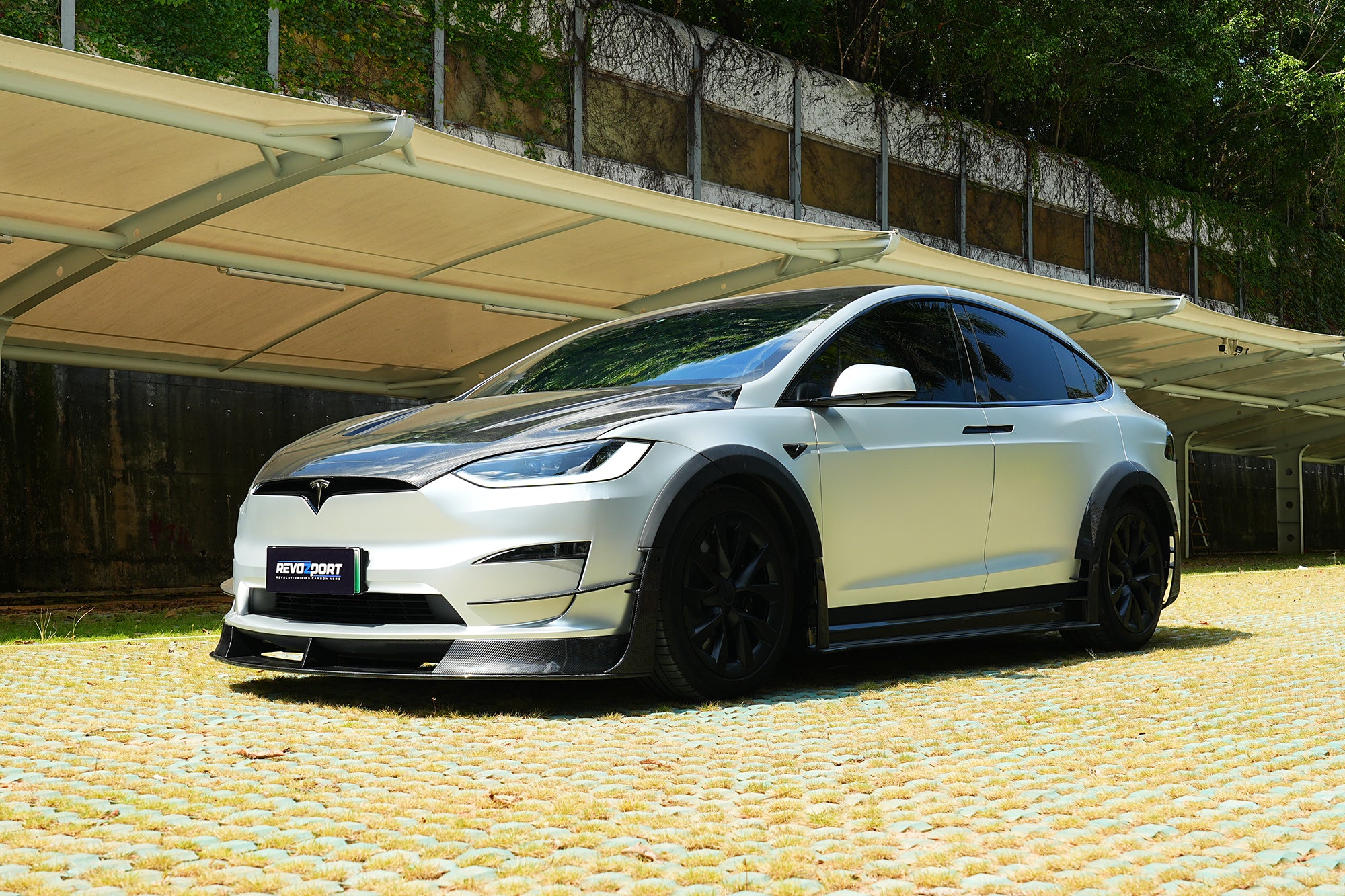 Silver Tesla Model X featuring a REVOZPORT body kit with carbon fiber front lip, side skirts, wide fender flares, and lowered stance parked outdoors in sunlight.