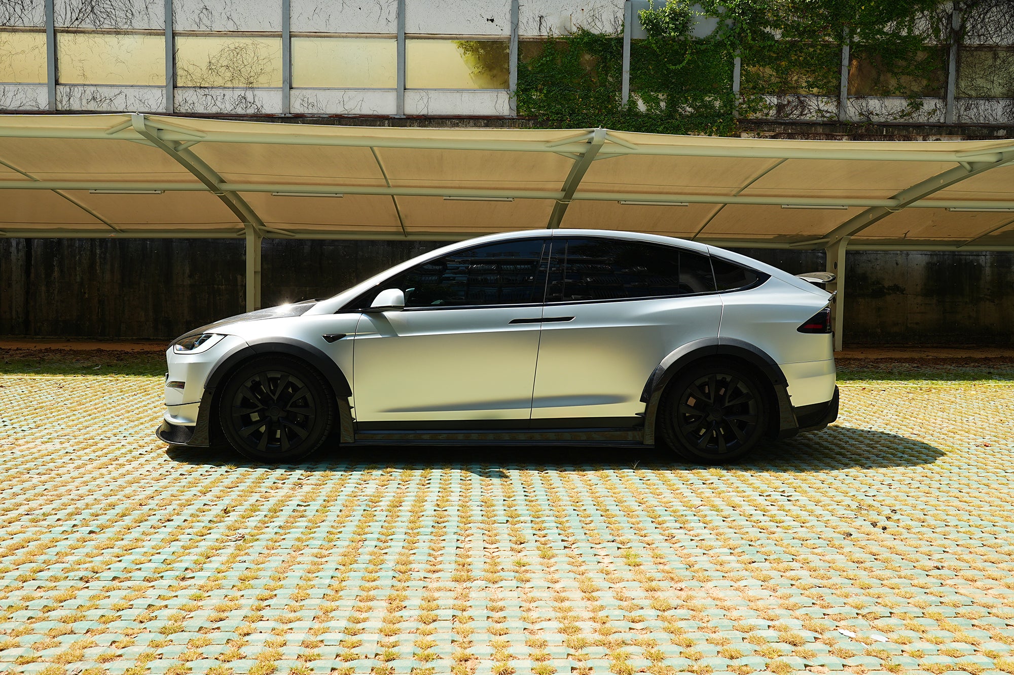 Silver Tesla Model X featuring a REVOZPORT body kit with carbon fiber front lip, side skirts, wide fender flares, and lowered stance parked outdoors in sunlight.