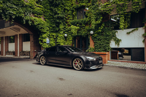 Matte black Audi with carbon fiber body kit and gold wheels parked beside ivy-covered brick architecture.