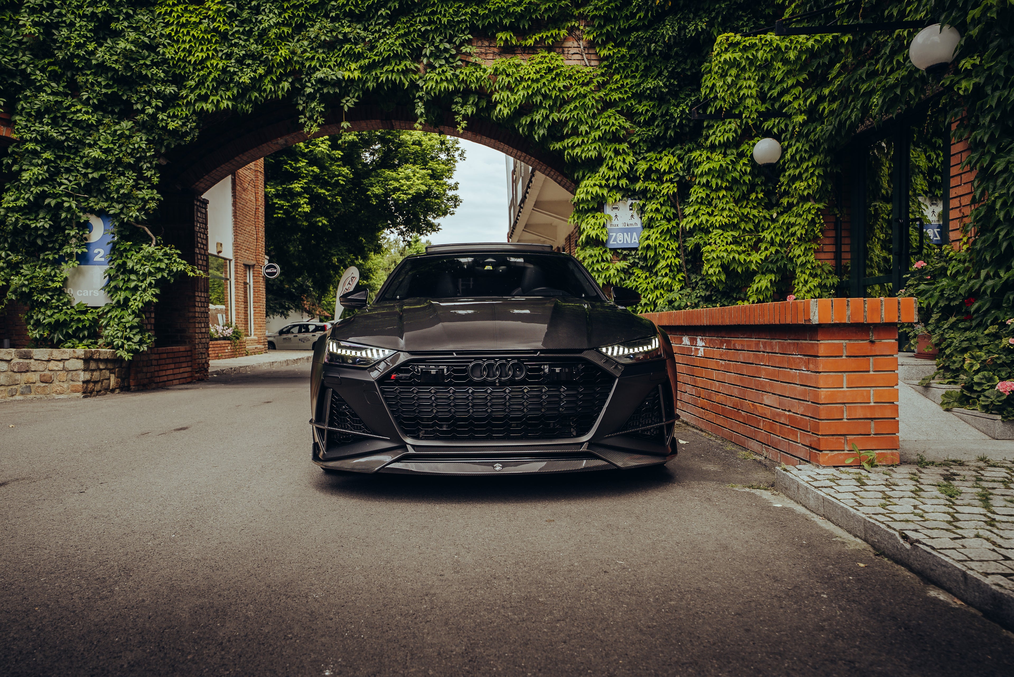 Black Audi with carbon fiber hood and front lip parked under an ivy-covered arch, showcasing aggressive front aero styling.