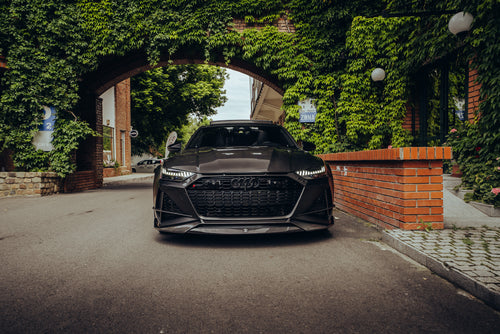 Black Audi with carbon fiber hood and front lip parked under an ivy-covered arch, showcasing aggressive front aero styling.
