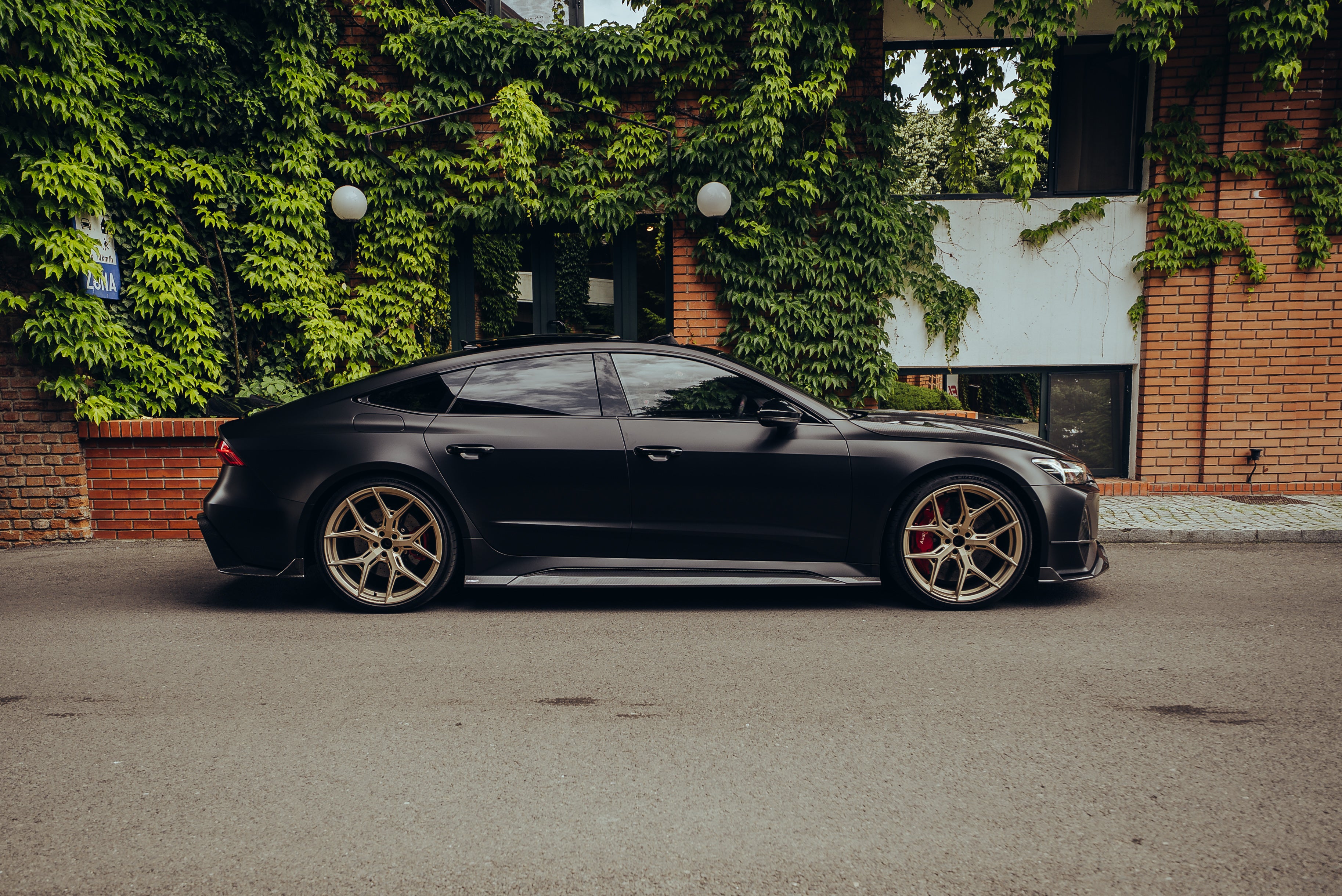 Side profile of a matte black Audi with carbon fiber body kit and gold wheels beside ivy-covered brick architecture.