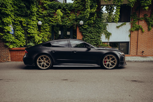 Side profile of a matte black Audi with carbon fiber body kit and gold wheels beside ivy-covered brick architecture.