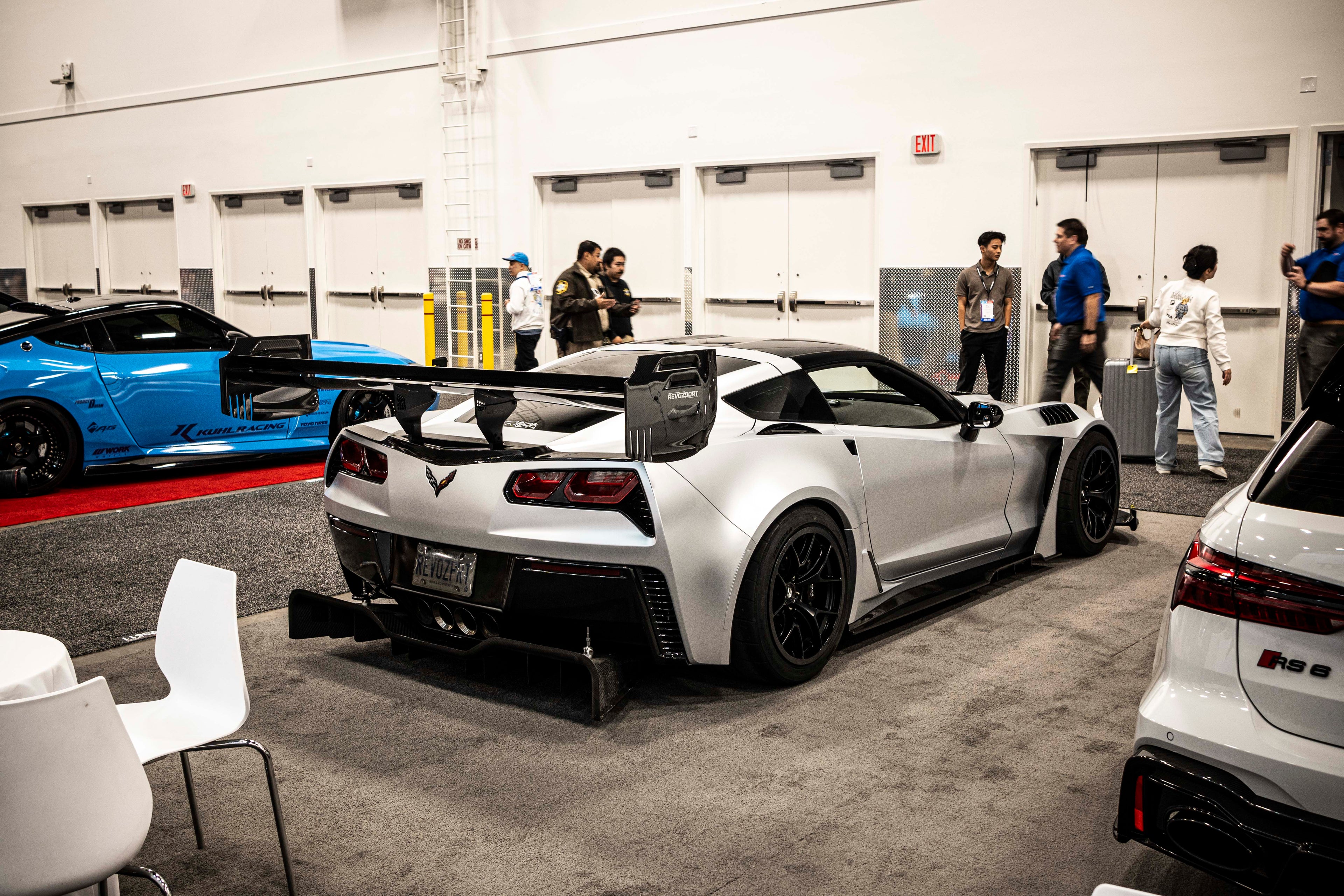 Silver Chevrolet Corvette race car displayed at an indoor automotive exhibition, featuring a wide-body aerodynamic kit, large rear wing, carbon fiber front splitter, and vented hood. The aggressive motorsport styling is showcased under exhibition lighting with REVOZPORT branding in the background.
