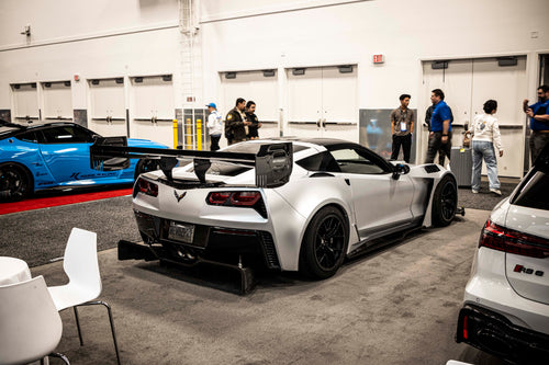 Silver Chevrolet Corvette race car displayed at an indoor automotive exhibition, featuring a wide-body aerodynamic kit, large rear wing, carbon fiber front splitter, and vented hood. The aggressive motorsport styling is showcased under exhibition lighting with REVOZPORT branding in the background.