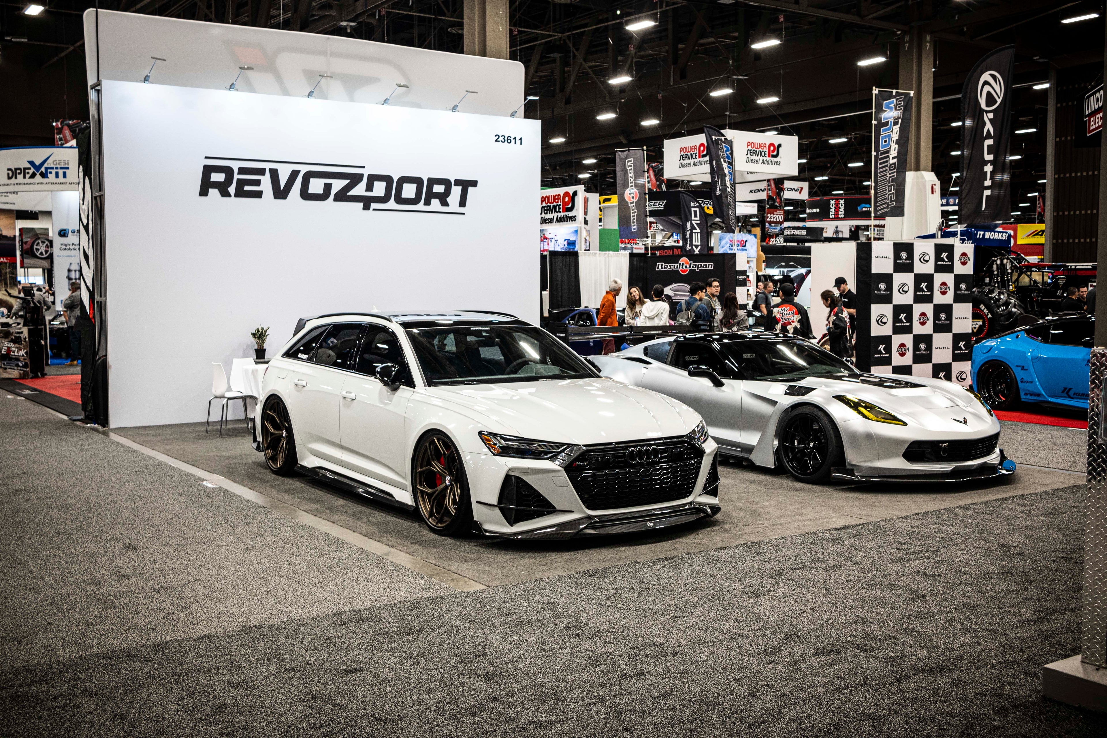 Silver Chevrolet Corvette race car displayed at an indoor automotive exhibition, featuring a wide-body aerodynamic kit, large rear wing, carbon fiber front splitter, and vented hood. The aggressive motorsport styling is showcased under exhibition lighting with REVOZPORT branding in the background.