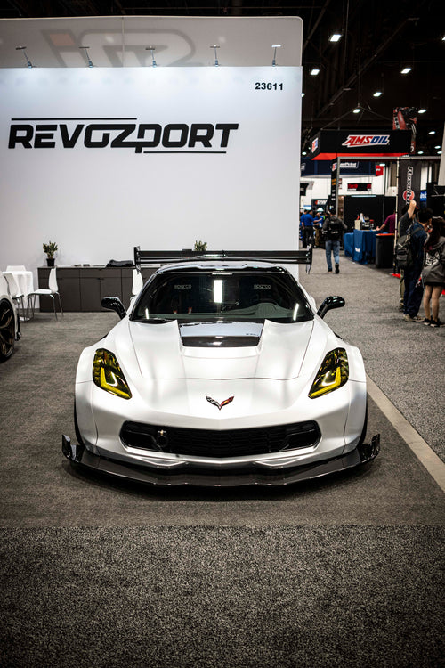 Silver Chevrolet Corvette race car displayed at an indoor automotive exhibition, featuring a wide-body aerodynamic kit, large rear wing, carbon fiber front splitter, and vented hood. The aggressive motorsport styling is showcased under exhibition lighting with REVOZPORT branding in the background.