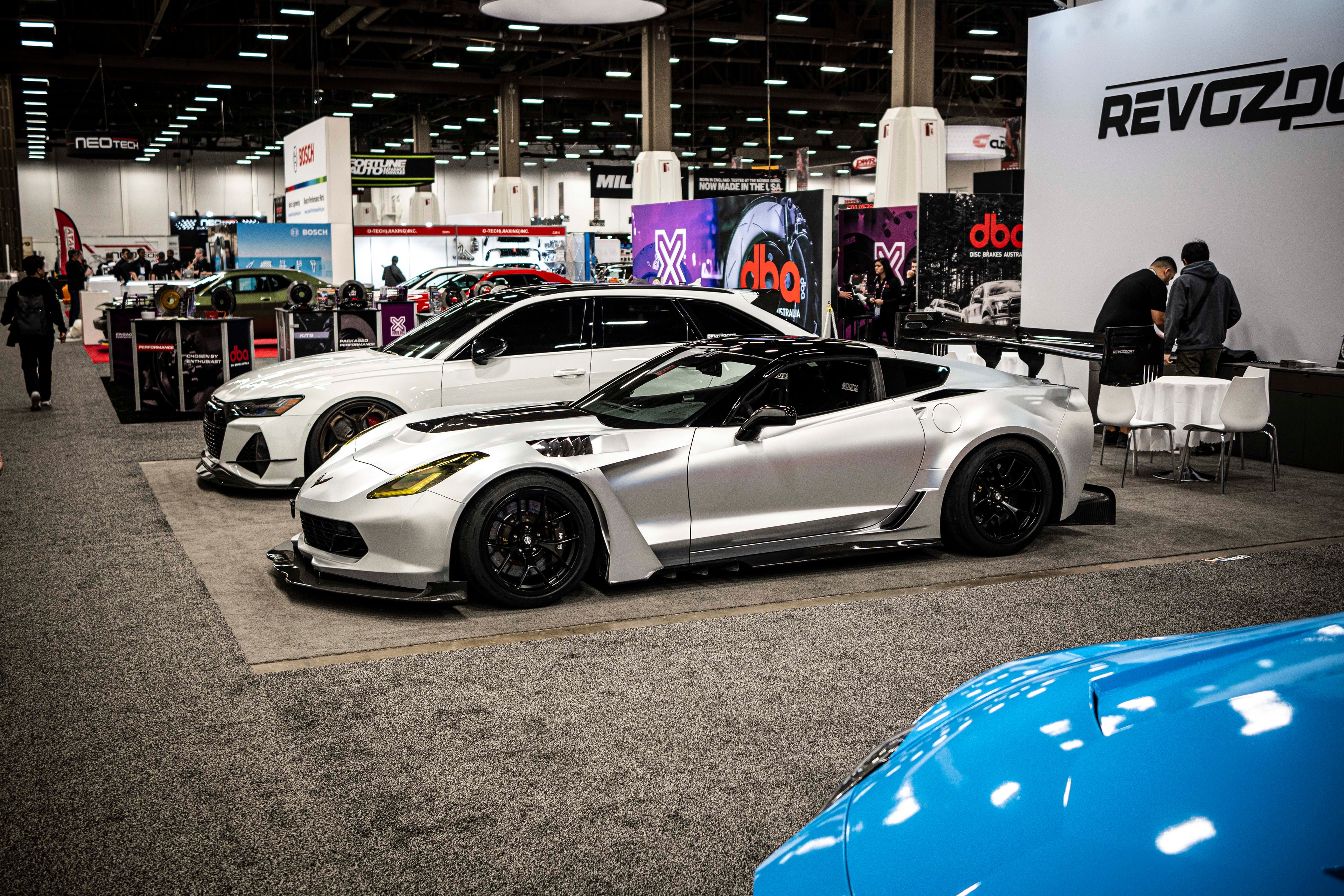 Silver Chevrolet Corvette race car displayed at an indoor automotive exhibition, featuring a wide-body aerodynamic kit, large rear wing, carbon fiber front splitter, and vented hood. The aggressive motorsport styling is showcased under exhibition lighting with REVOZPORT branding in the background.