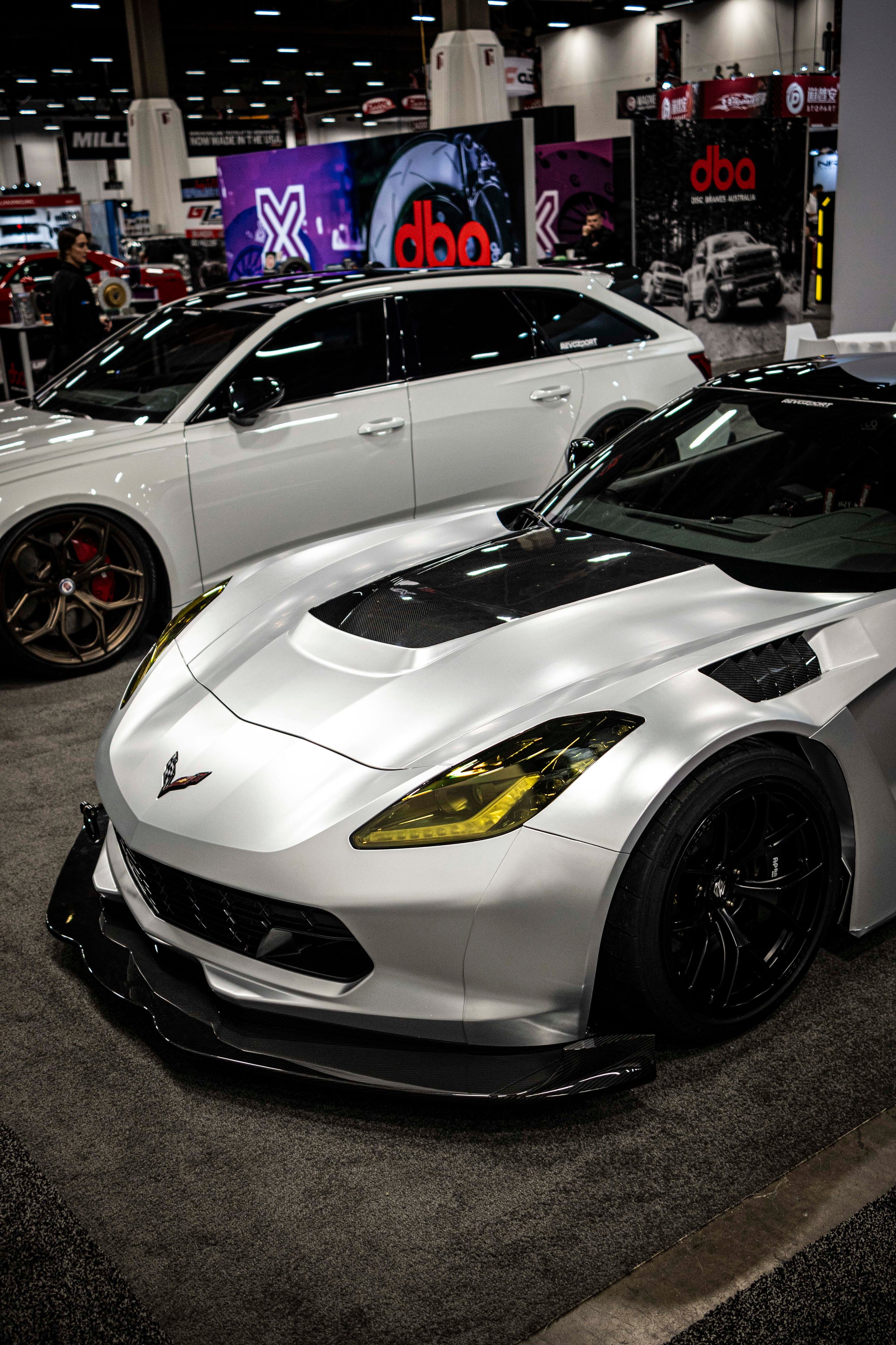 Silver Chevrolet Corvette race car displayed at an indoor automotive exhibition, featuring a wide-body aerodynamic kit, large rear wing, carbon fiber front splitter, and vented hood. The aggressive motorsport styling is showcased under exhibition lighting with REVOZPORT branding in the background.