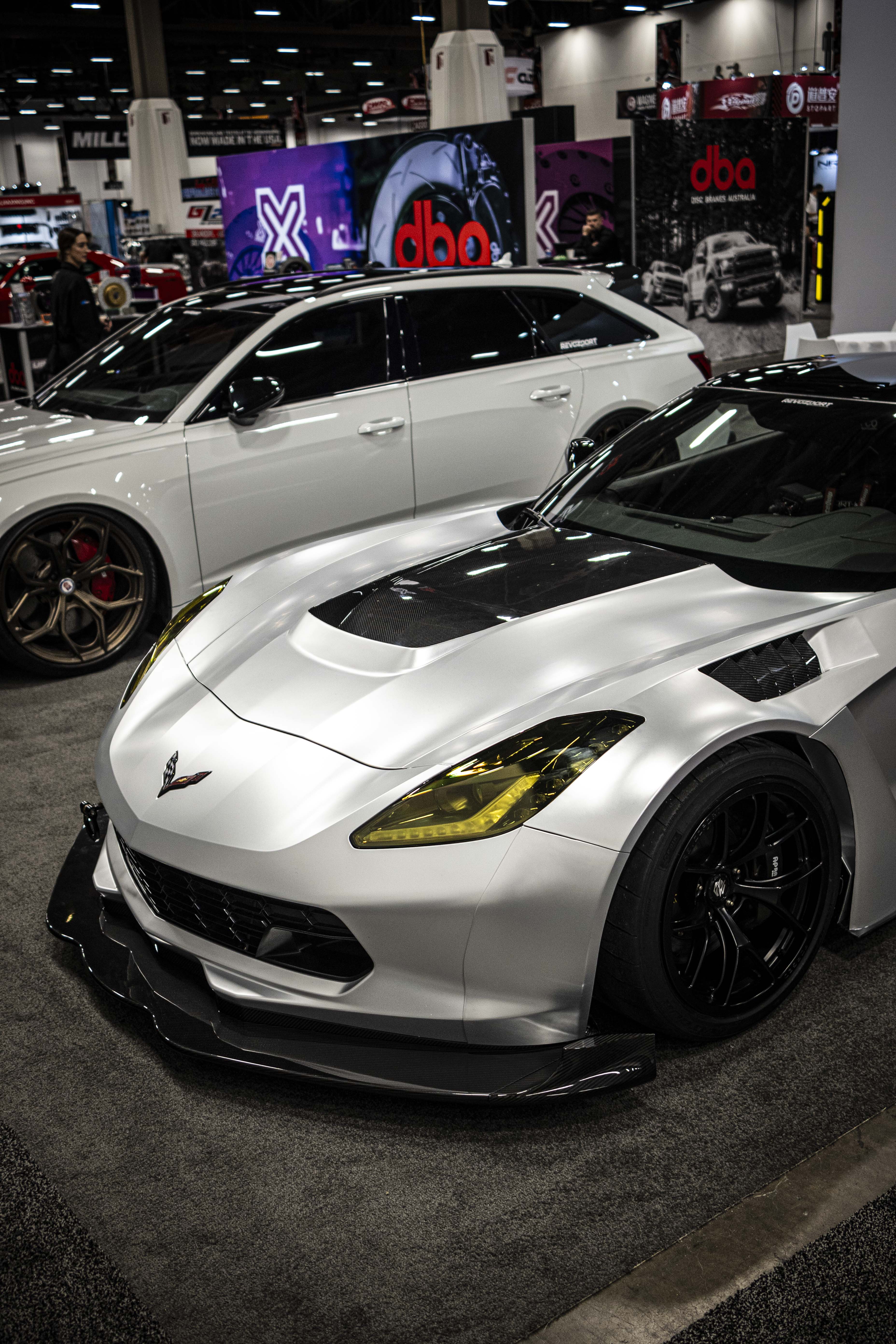 Silver Chevrolet Corvette race car displayed at an indoor automotive exhibition, featuring a wide-body aerodynamic kit, large rear wing, carbon fiber front splitter, and vented hood. The aggressive motorsport styling is showcased under exhibition lighting with REVOZPORT branding in the background.