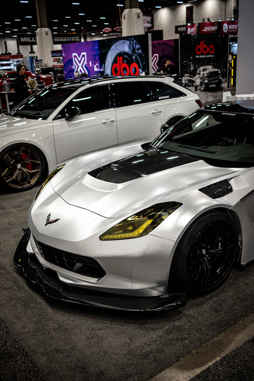 Silver Chevrolet Corvette race car displayed at an indoor automotive exhibition, featuring a wide-body aerodynamic kit, large rear wing, carbon fiber front splitter, and vented hood. The aggressive motorsport styling is showcased under exhibition lighting with REVOZPORT branding in the background.