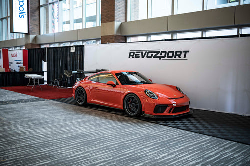 Close-up of a performance wheel setup on an orange Porsche sports car, featuring black multi-spoke forged rims, red brake calipers, and a large drilled brake rotor. The image highlights the aggressive fitment, low-profile tire, and carbon fiber side skirt details at an indoor automotive display.