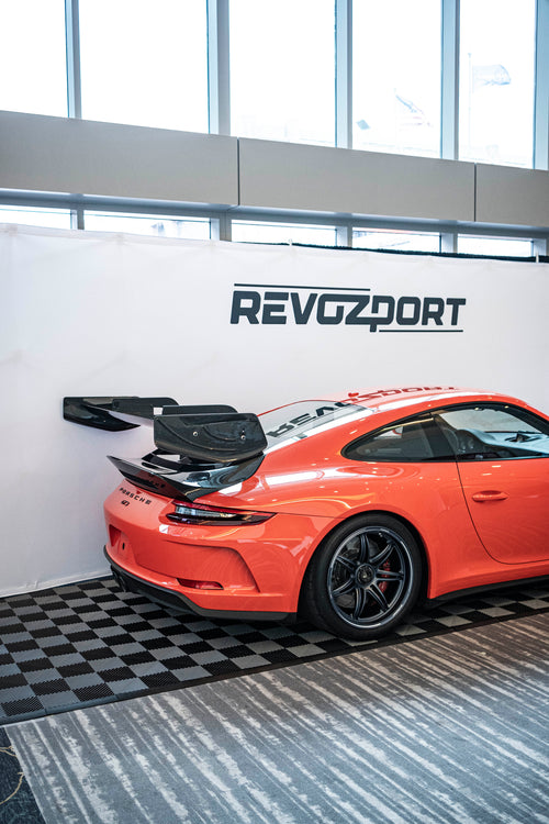 Close-up of a performance wheel setup on an orange Porsche sports car, featuring black multi-spoke forged rims, red brake calipers, and a large drilled brake rotor. The image highlights the aggressive fitment, low-profile tire, and carbon fiber side skirt details at an indoor automotive display.