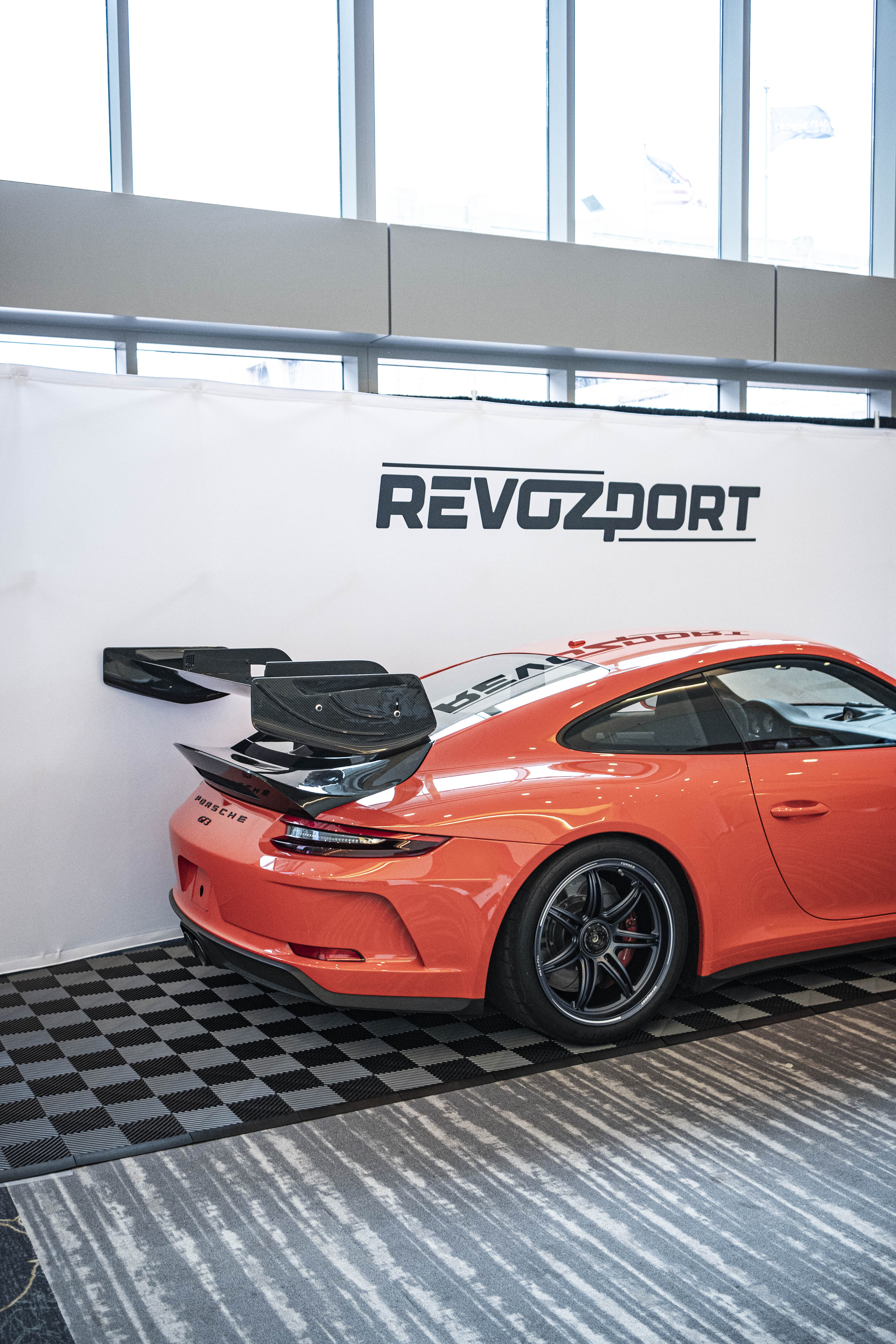 An orange Porsche sports car displayed indoors at an automotive exhibition booth, featuring an aggressive front splitter, black performance wheels, and a sleek aerodynamic body kit. The car is parked in front of a REVOZPORT branded display wall inside a modern convention center with large windows and exhibition setups in the background.