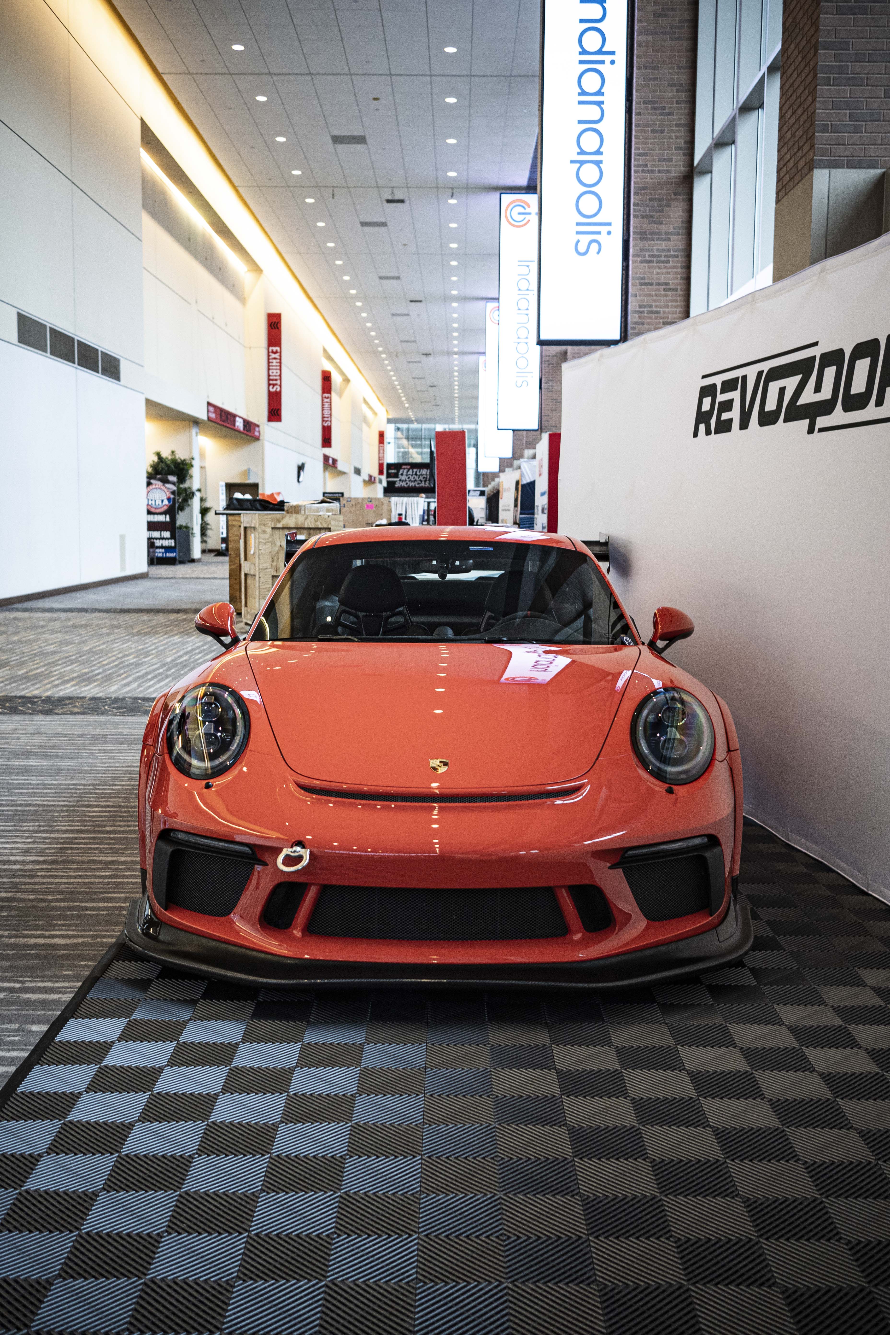 An orange Porsche sports car displayed indoors at an automotive exhibition booth, featuring an aggressive front splitter, black performance wheels, and a sleek aerodynamic body kit. The car is parked in front of a REVOZPORT branded display wall inside a modern convention center with large windows and exhibition setups in the background.