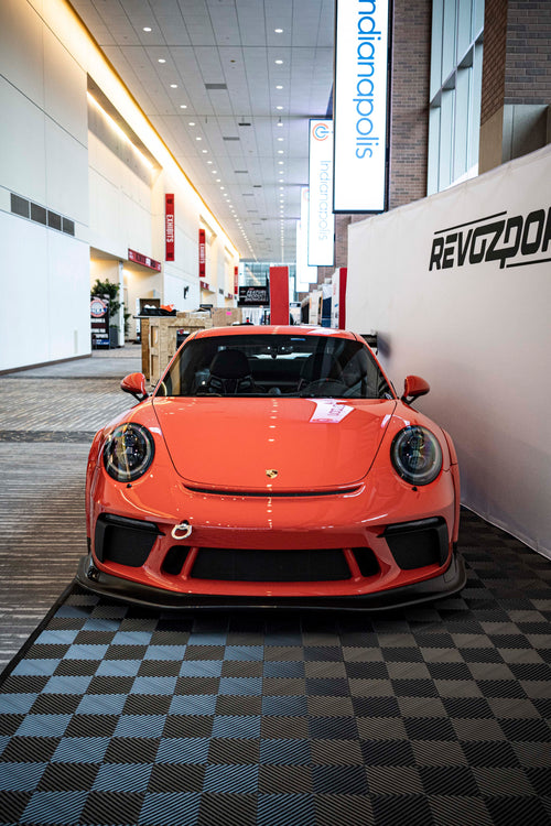 An orange Porsche sports car displayed indoors at an automotive exhibition booth, featuring an aggressive front splitter, black performance wheels, and a sleek aerodynamic body kit. The car is parked in front of a REVOZPORT branded display wall inside a modern convention center with large windows and exhibition setups in the background.
